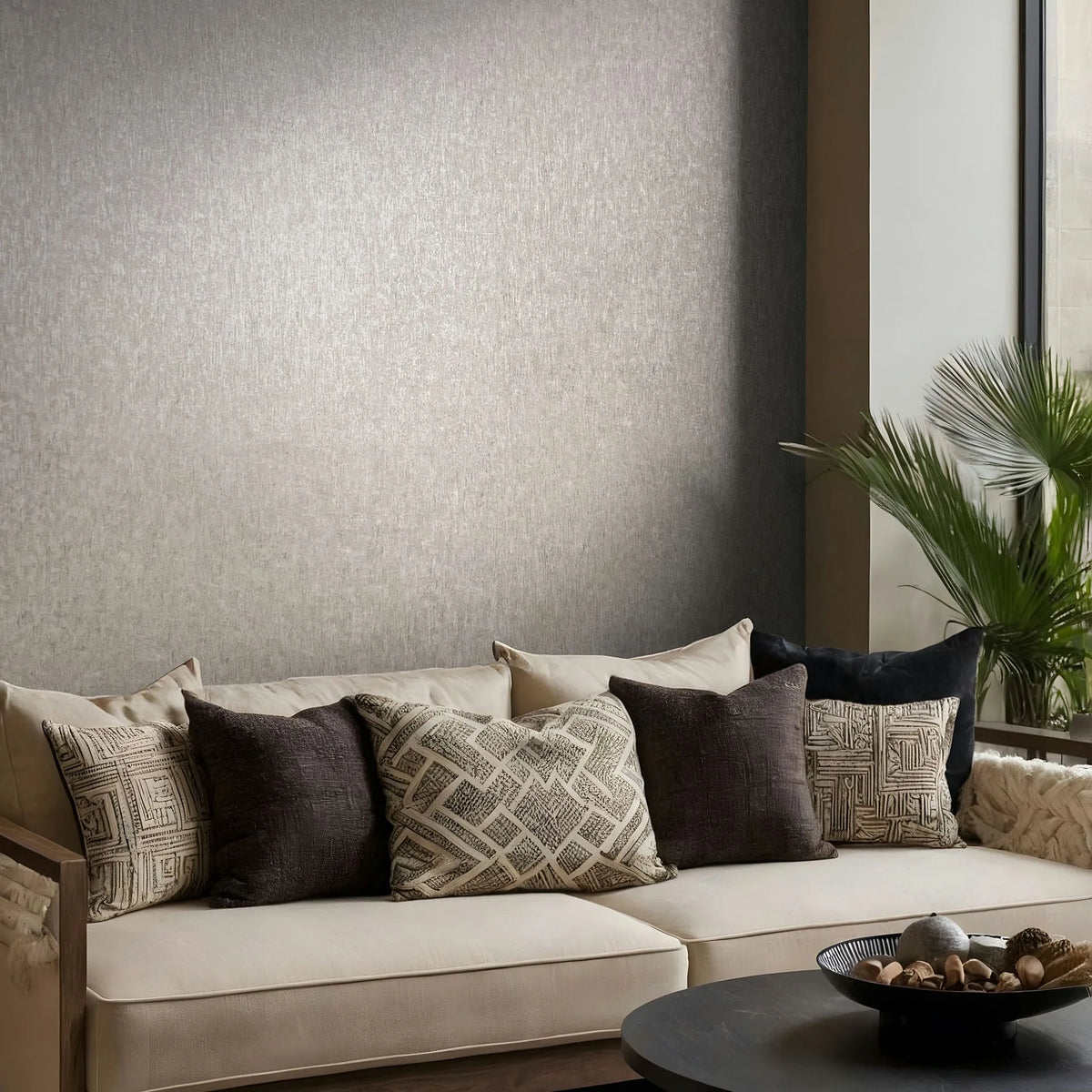 Marburg Ritus 35525 Textured gray wallpaper on a living room wall behind a beige sofa with patterned cushions, a palm plant by the window, and a round coffee table with a decorative bowl.