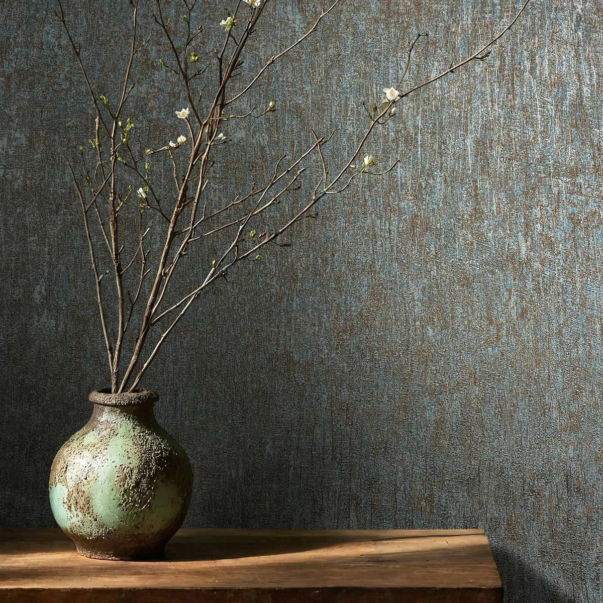 Marburg Ritus 35526 Textured blue and brown wallpaper shown behind a rustic ceramic vase with flowering branches on a wooden surface.