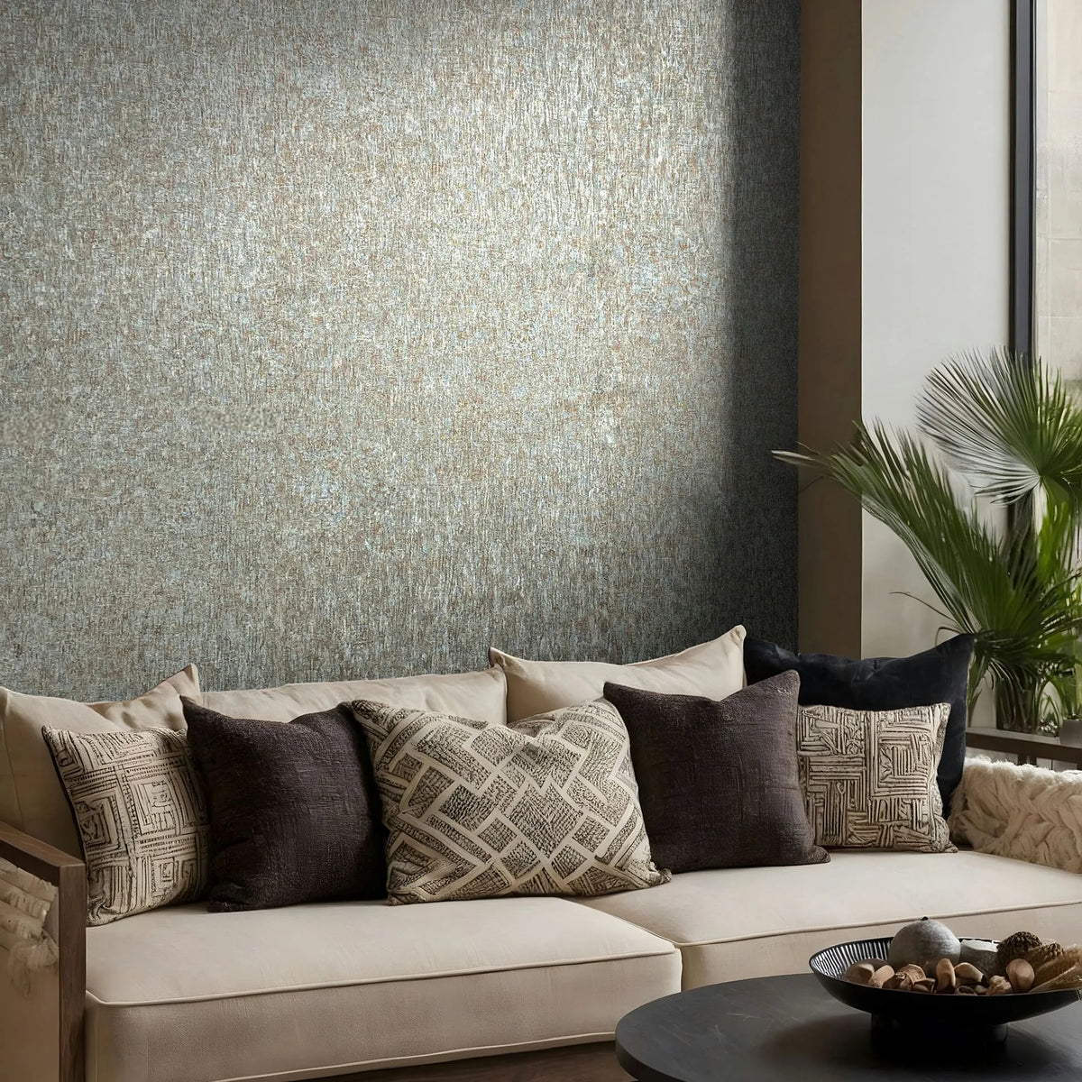 Marburg Ritus 35526 Textured blue and copper wallpaper on a feature wall behind a cream sofa with patterned cushions, a round coffee table with a bowl, and a leafy plant by a tall window.