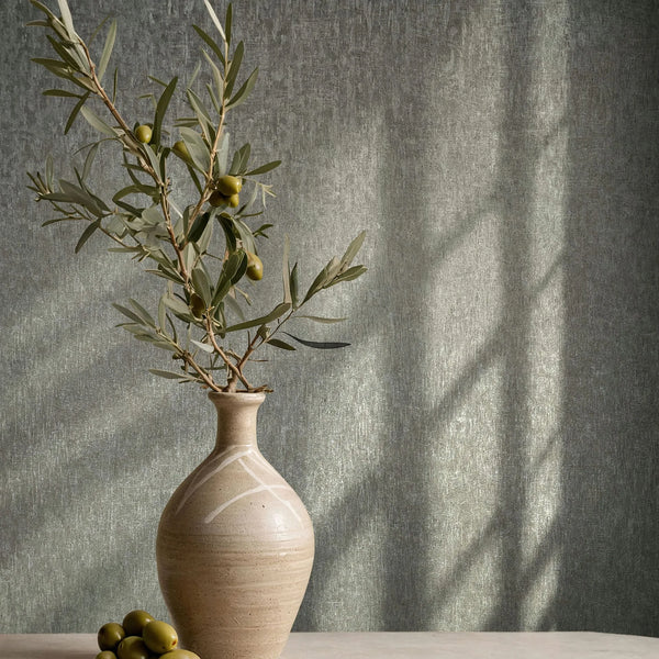Marburg Ritus 35528 Textured gray wallpaper with a fine mottled strie, shown behind a ceramic vase holding an olive branch with a few olives on a tabletop; soft windowpane shadows fall across the wall.
