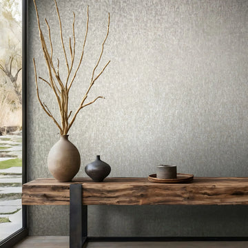 Marburg Ritus 35528 Subtle gray mottled-texture wallpaper shown behind a wood console table with a tall vase of branches, a small vase, and a tray; a window at left reveals a garden view.