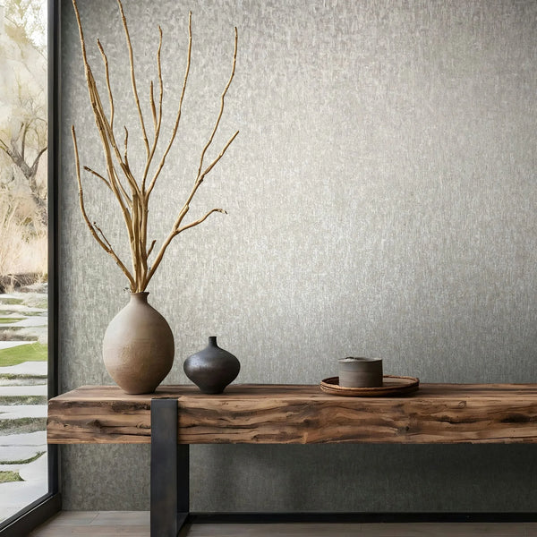 Marburg Ritus 35528 Subtle gray mottled-texture wallpaper shown behind a wood console table with a tall vase of branches, a small vase, and a tray; a window at left reveals a garden view.