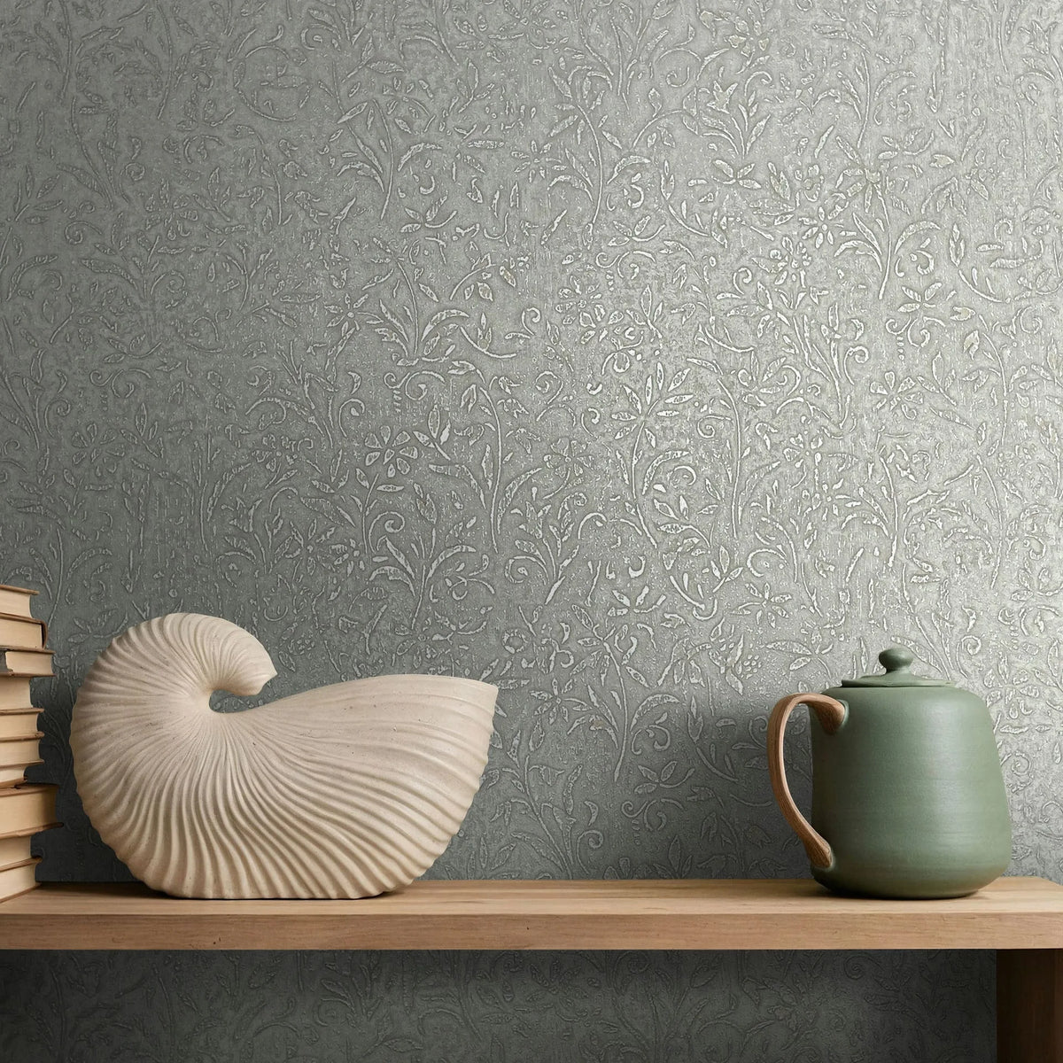 Marburg Ritus 35529 Gray wallpaper with an embossed floral vine pattern behind a wooden shelf holding stacked books, a shell-shaped sculpture, and a green teapot.