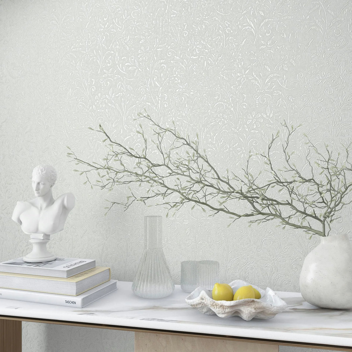 Marburg Ritus 35530 White embossed wallpaper with delicate botanical vines behind a styled console featuring a bust sculpture, books, glass vessels, a white vase with branches, and a shell bowl holding lemons on a marble surface.