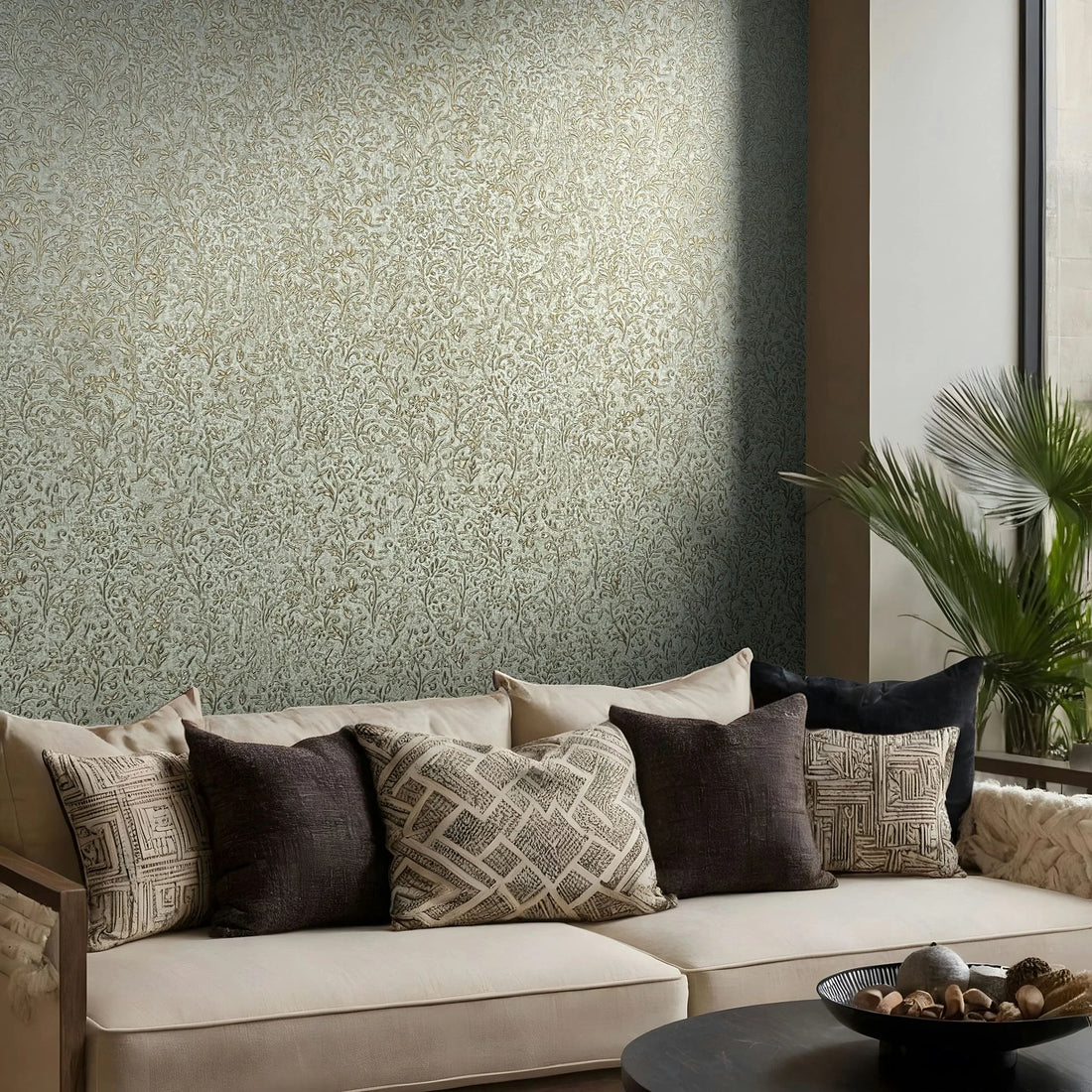 Marburg Ritus 35532 Living room scene with blue wallpaper featuring a gold floral vine pattern behind a beige sofa with patterned cushions, a coffee table with a decorative bowl, and a leafy plant beside a tall window.