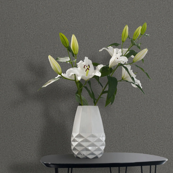 Marburg Ritus 35541 Styled detail of medium-dark gray textured wallpaper behind a white faceted vase holding white lilies on a small black table.