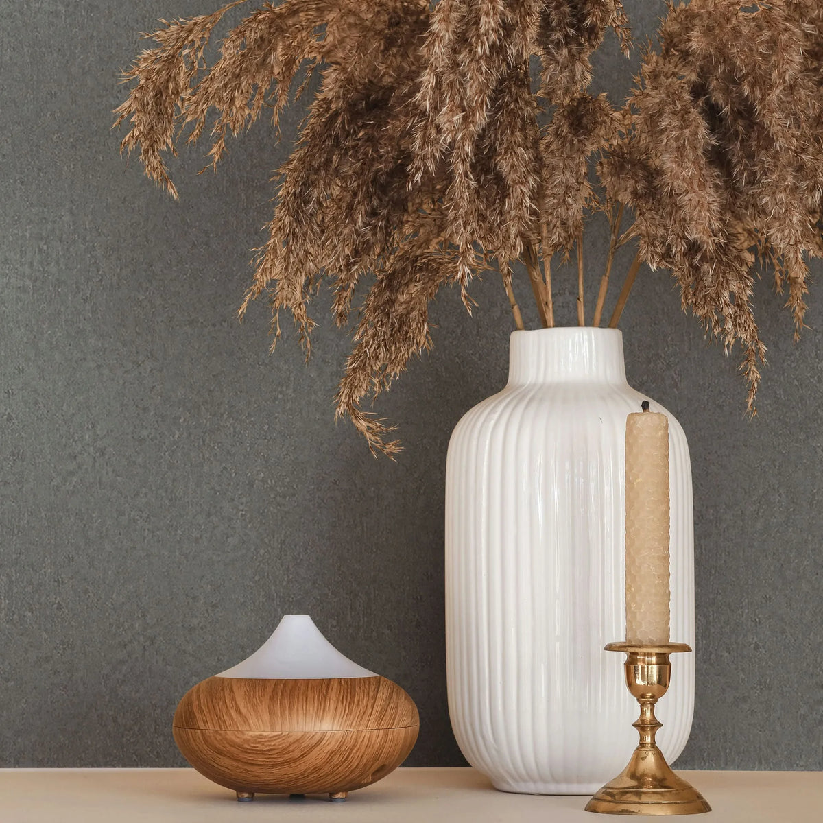 Marburg Ritus 35541 Gray textured wallpaper behind a white ribbed vase filled with dried grasses, a small wood-and-white diffuser, and a brass candlestick on a light surface.