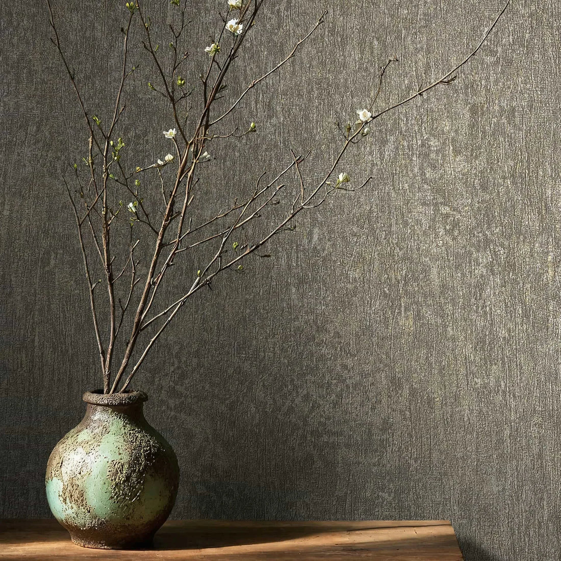 Marburg Ritus 35552 Close-up of brown‑gray textured wallpaper behind a rustic ceramic vase holding bare branches with small white blossoms on a wooden shelf.