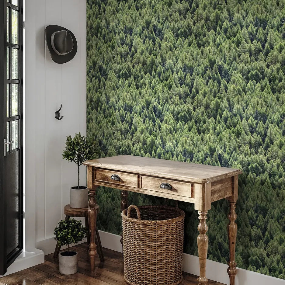 Marburg Struktura 45730 Green forest treetops wallpaper behind a wooden console table with a wicker basket; potted plants, a wall hook with a hat, and a door are visible.