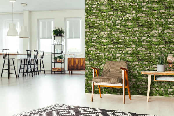 Marburg Struktura 45732 Green mossy stone brick effect wallpaper on a feature wall beside a wood armchair and console table, with plants, a table lamp, rug, shelving, bar stools, pendant lights, and windows in an open-plan room.