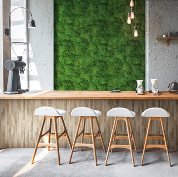 Marburg Struktura 45733 Green moss-texture wallpaper used as a feature wall behind a wood bar counter with four white-seated stools, pendant bulbs, a coffee grinder, pour-over drippers, and a kettle.