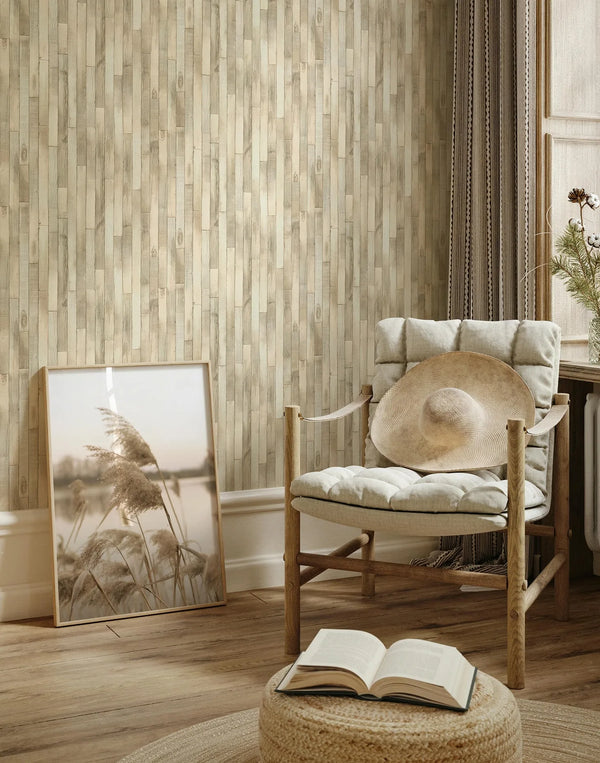 Marburg Struktura 45734 Beige vertical wood plank–effect wallpaper in a cozy living room corner with a cushioned wooden chair holding a sun hat, a framed print leaning against the wall, a pouf with an open book, and a window with curtains.