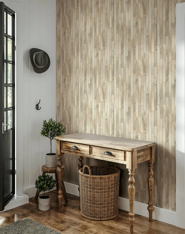 Marburg Struktura 45734 Beige wallpaper with a vertical wood‑plank effect is shown on an entry wall behind a wood console table, with a wicker basket below, two potted plants on a stool and the floor, a wall hook with a hat, and a glass‑paneled door.