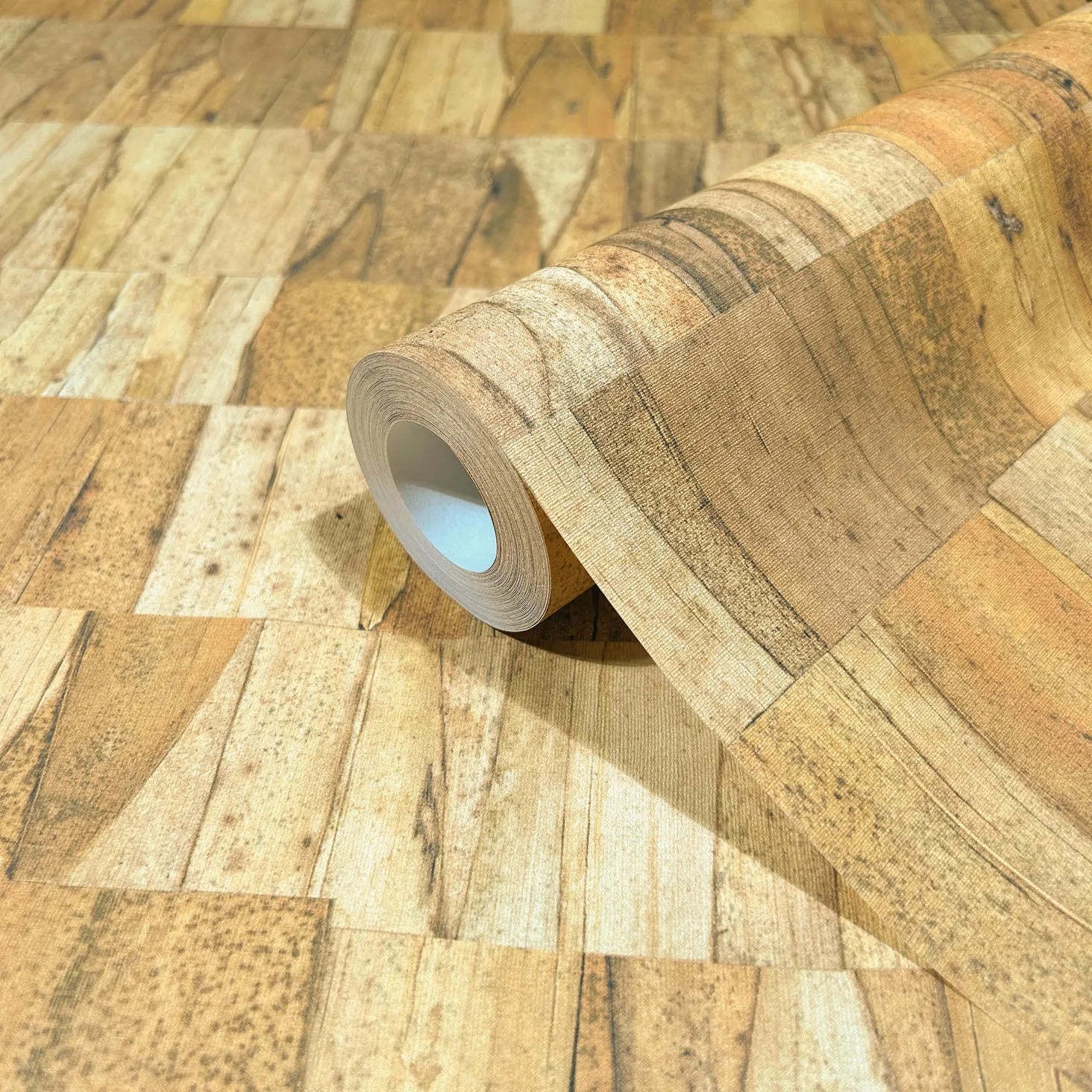 Marburg Struktura 45737 Close-up of a wallpaper roll resting on matching faux wood plank patchwork in warm beige and brown tones, showing the printed grain and rectangular segments.