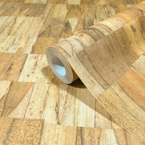 Marburg Struktura 45737 Close-up of a wallpaper roll resting on matching faux wood plank patchwork in warm beige and brown tones, showing the printed grain and rectangular segments.