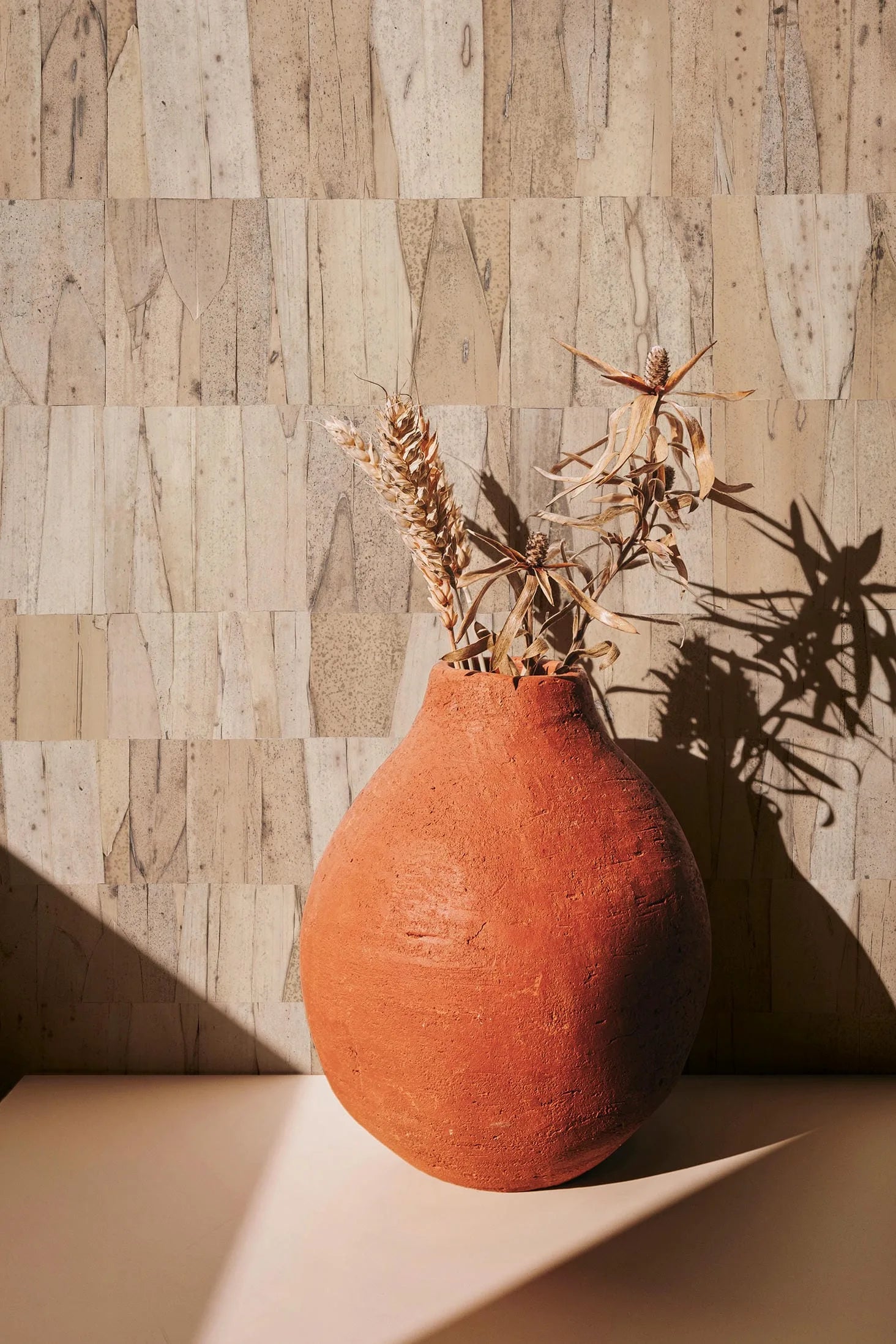 Marburg Struktura 45738 Light brown patchwork wallpaper made of narrow rectangular panels with subtle organic grain. A terracotta clay vase holding dried grasses sits on a light surface with strong sunlight casting shadows.