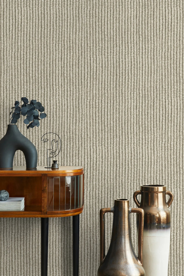 Marburg Struktura 45748 Beige and gray wallpaper with narrow vertical braided chevron lines, shown behind a wooden console with a sculptural dark vase holding leafy stems, a small wire-face ornament, and two tall handled vases.