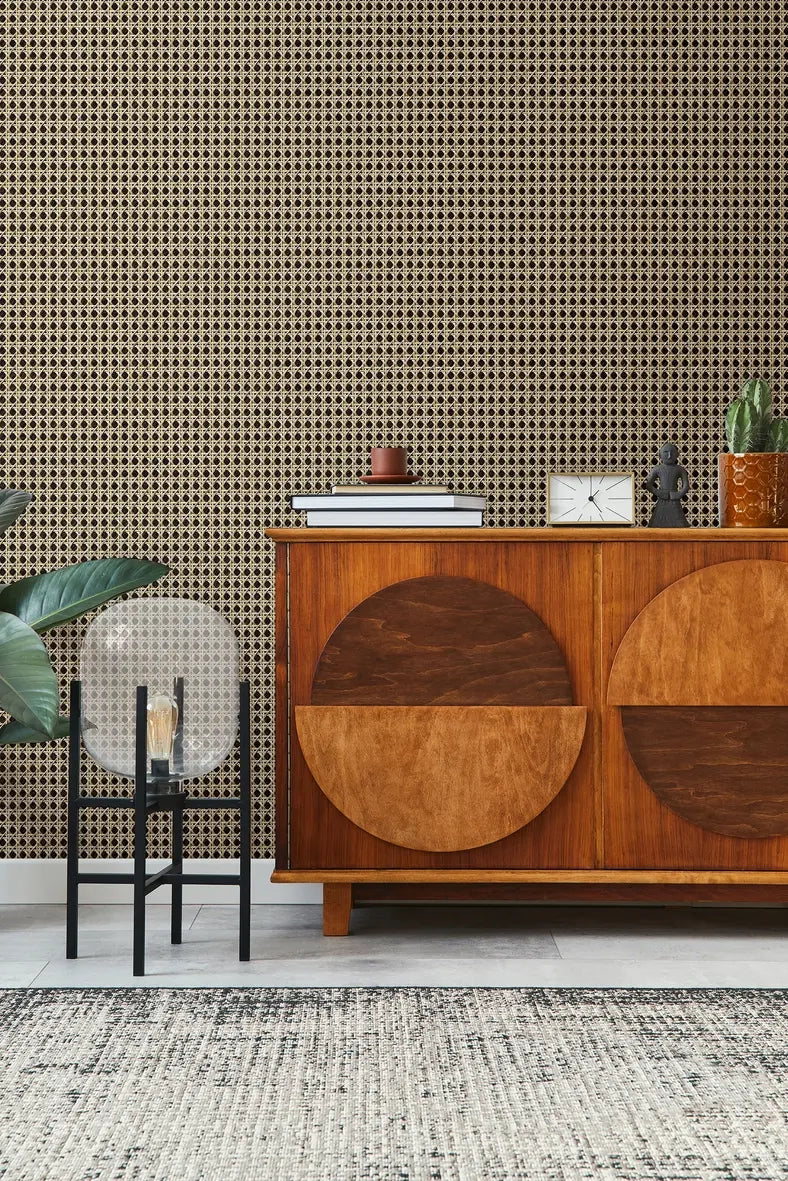 Marburg Struktura 45751 Beige and brown geometric lattice wallpaper behind a mid-century wood sideboard with stacked books, a small cup, a clock, a figurine, a potted plant, and a glass table lamp, with a rug in the foreground.