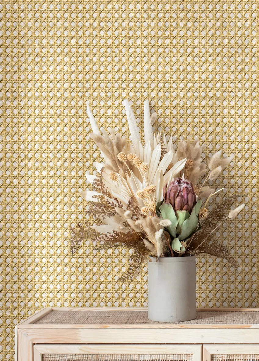 Marburg Struktura 45753 Geometric wallpaper with a small repeating lattice in warm yellow-gold on a light background, shown behind a light wood cabinet with a cylindrical vase of dried flowers.