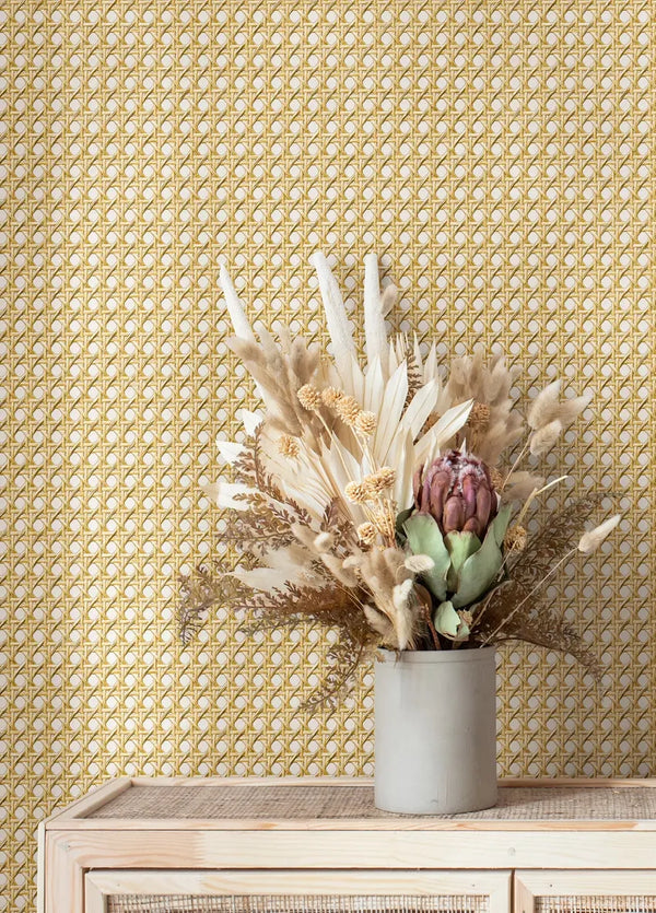 Marburg Struktura 45753 Geometric wallpaper with a small repeating lattice in warm yellow-gold on a light background, shown behind a light wood cabinet with a cylindrical vase of dried flowers.