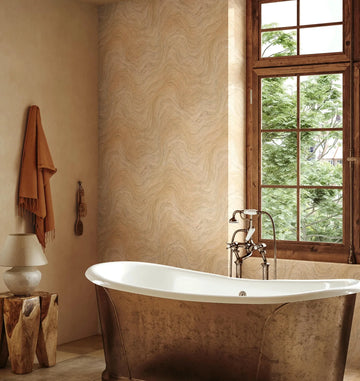 Marburg Struktura 45755 Bathroom scene featuring a beige wavy-pattern wallpaper on a feature wall beside a wood-framed window. A freestanding tub with exposed faucet sits in front, with a hanging towel and bath brushes, a small lamp, and a wood stump side table visible.