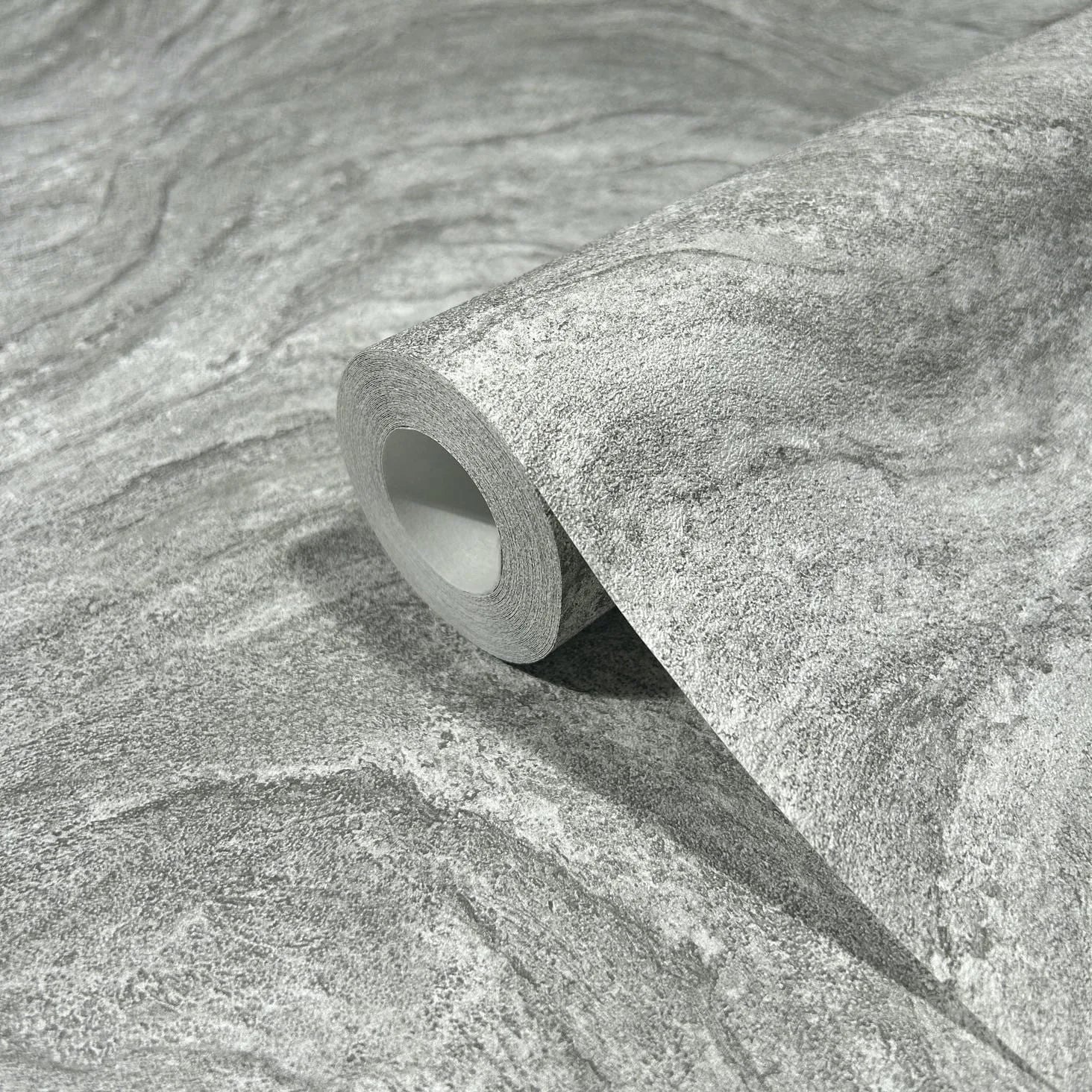 Marburg Struktura 45758 Close-up of a gray stone-effect wallpaper roll resting on a matching unrolled sheet, showing fine grain and gentle wavy veining.