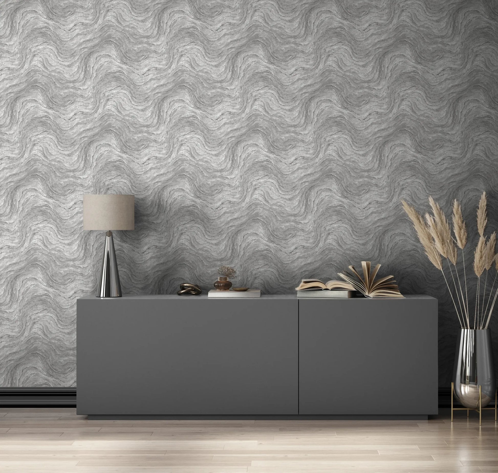 Marburg Struktura 45758 Gray wallpaper with a tonal wavy, marbled pattern behind a low modern sideboard. A table lamp, small decor pieces, stacked and open books, and a tall glass vase with pampas stems sit on the cabinet, with light wood flooring below.