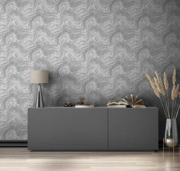 Marburg Struktura 45758 Gray wallpaper with a tonal wavy, marbled pattern behind a low modern sideboard. A table lamp, small decor pieces, stacked and open books, and a tall glass vase with pampas stems sit on the cabinet, with light wood flooring below.