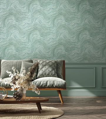 Marburg Struktura 45759 Green wallpaper with a flowing wavy, marbled texture shown in a living room above painted panel moulding, with a cushioned wooden-frame sofa, patterned pillows, a low wooden coffee table, a vase of dried stems, a round woven rug, and herringbone wood flooring.