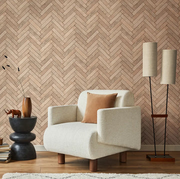 Marburg Struktura 45760 Beige herringbone wallpaper shown in a styled room with a cream armchair, brown cushion, stacked side table with books, a wooden animal figurine and a vase with stems, a tall two-shade floor lamp, wood floor, and a soft rug.