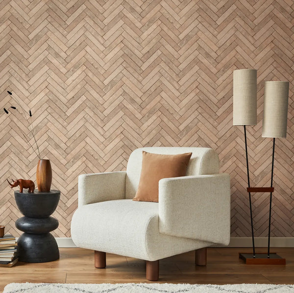 Marburg Struktura 45760 Beige herringbone wallpaper shown in a styled room with a cream armchair, brown cushion, stacked side table with books, a wooden animal figurine and a vase with stems, a tall two-shade floor lamp, wood floor, and a soft rug.
