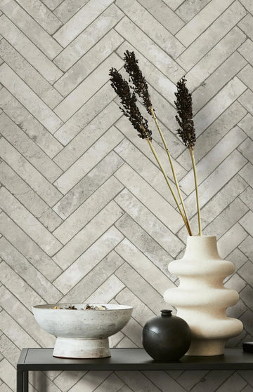 Marburg Struktura 45762 Gray stone-look herringbone wallpaper behind a dark shelf holding a sculptural light-colored vase with dried stems, a round black vase, and a white bowl.