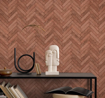 Marburg Struktura 45763 Reddish-brown herringbone brick-look wallpaper behind a black console shelf with an abstract stone sculpture, circular metal decor, and books including an open book.