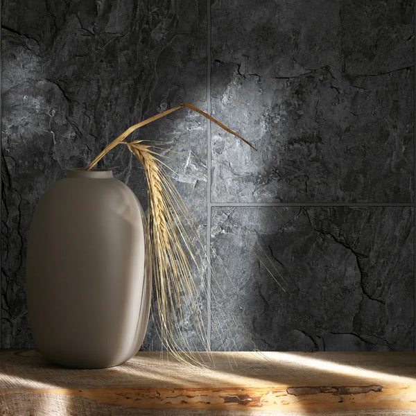 Marburg Struktura 45765 Dark grey stone-slab effect wallpaper with fine panel lines, shown behind a matte ceramic vase with a dried grass stem on a wood shelf under angled light.