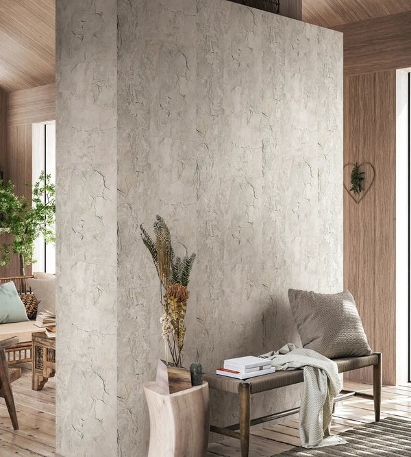 Marburg Struktura 45767 Light gray stone-effect wallpaper covers a central partition wall in a living room. A bench with a cushion and throw sits beside a floor vase holding dried foliage, with a rug and indoor greenery nearby.