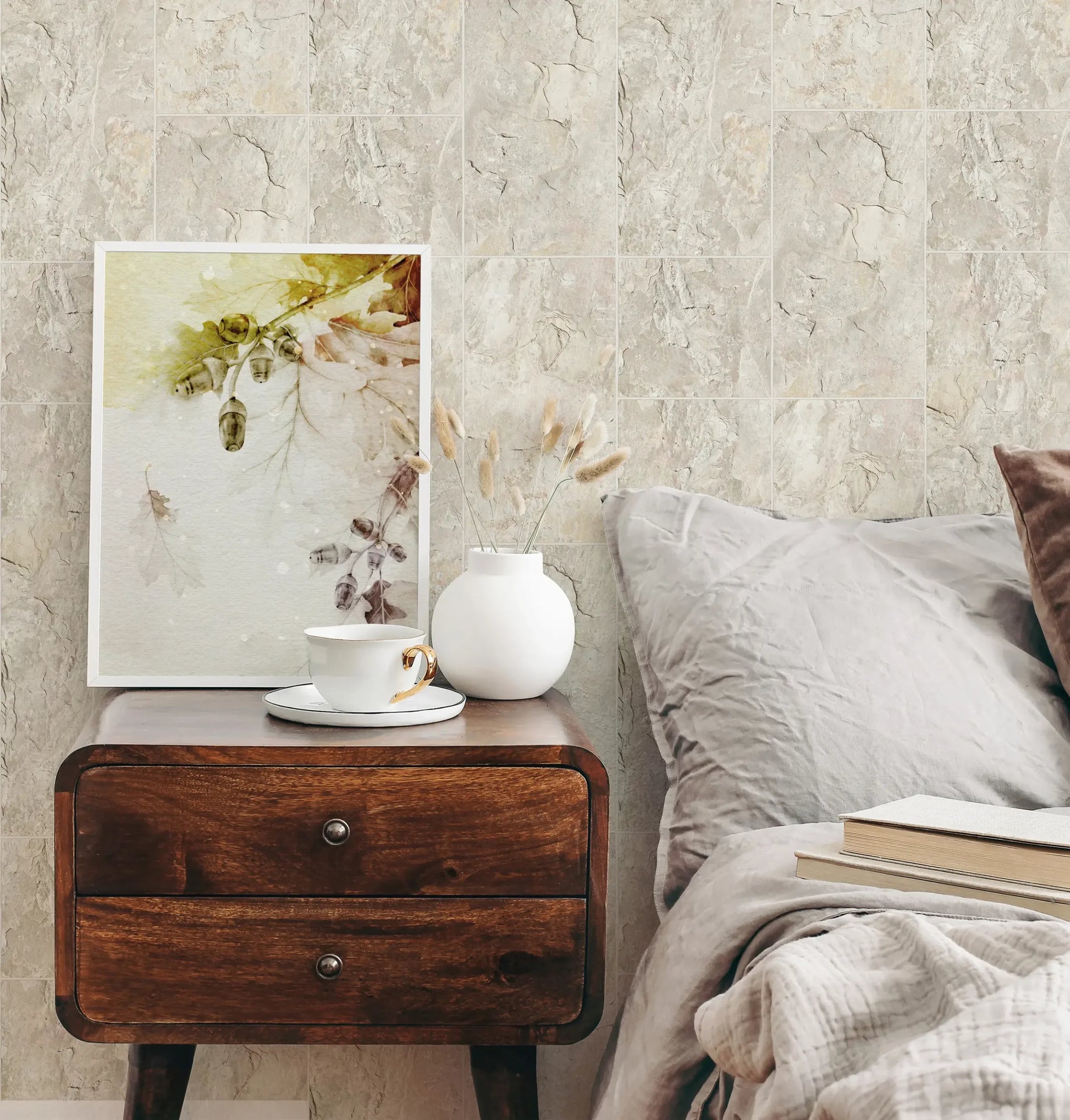 Marburg Struktura 45768 Bedroom scene with beige stone‑look tiled wallpaper behind a wooden nightstand holding a framed botanical print, a white vase with dried stems, and a cup and saucer beside a bed with pillows and books.