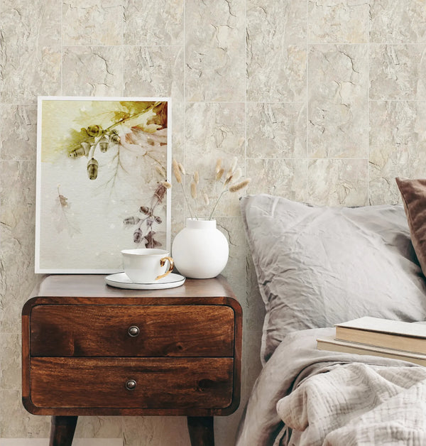 Marburg Struktura 45768 Bedroom scene with beige stone‑look tiled wallpaper behind a wooden nightstand holding a framed botanical print, a white vase with dried stems, and a cup and saucer beside a bed with pillows and books.