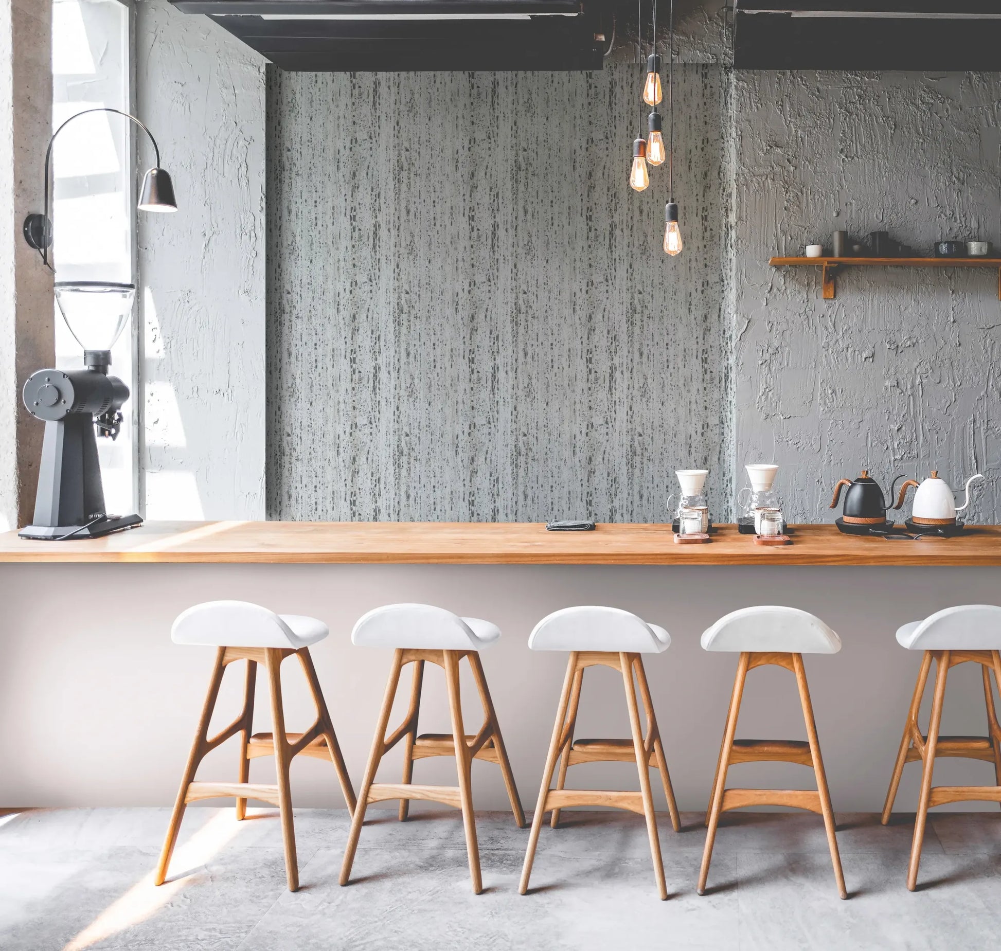 Marburg Travertino 33350 Cafe-style bar scene featuring a light gray, vertically streaked wallpaper as a feature wall behind a wooden counter with bar stools, pendant bulbs, a coffee grinder, pour-over drippers, and kettles.