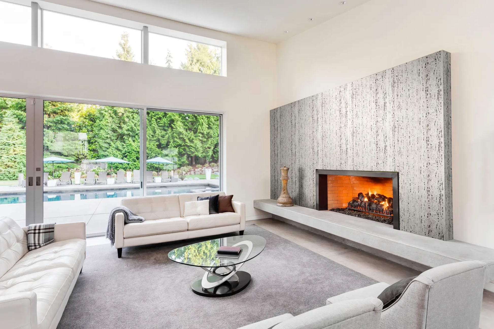 Marburg Travertino 33350 Vertical beige and gray striated wallpaper covers the fireplace wall in a bright modern living room with sofas, a glass coffee table, and a lit fireplace beside large windows overlooking a pool and trees.