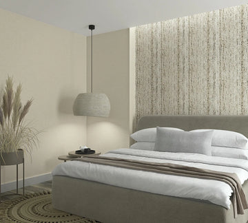 Marburg Travertino 33351 Bedroom scene featuring a vertical beige and taupe streaked wallpaper used as a feature wall behind the bed, with a coordinating solid light‑beige wall to the side. In front are an upholstered bed with white bedding and a gray pillow, a hanging pendant light,a.