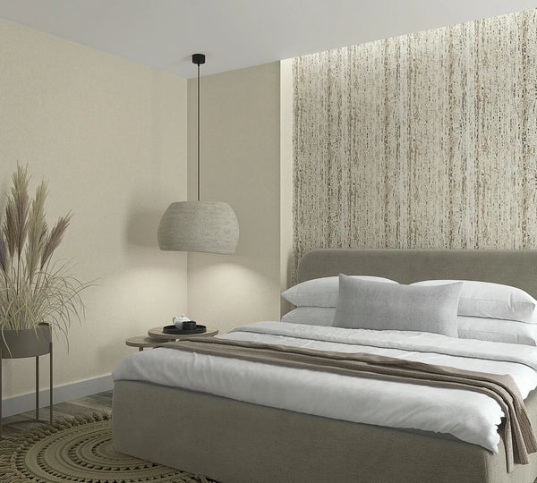 Marburg Travertino 33351 Bedroom scene featuring a vertical beige and taupe streaked wallpaper used as a feature wall behind the bed, with a coordinating solid light‑beige wall to the side. In front are an upholstered bed with white bedding and a gray pillow, a hanging pendant light,a.