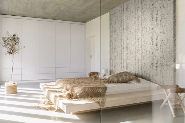 Marburg Travertino 33351 Beige, vertically striated wallpaper forms a feature wall behind a low platform bed in a minimalist bedroom with white built-in wardrobes, a potted tree, a bedside lamp, and a small stool.