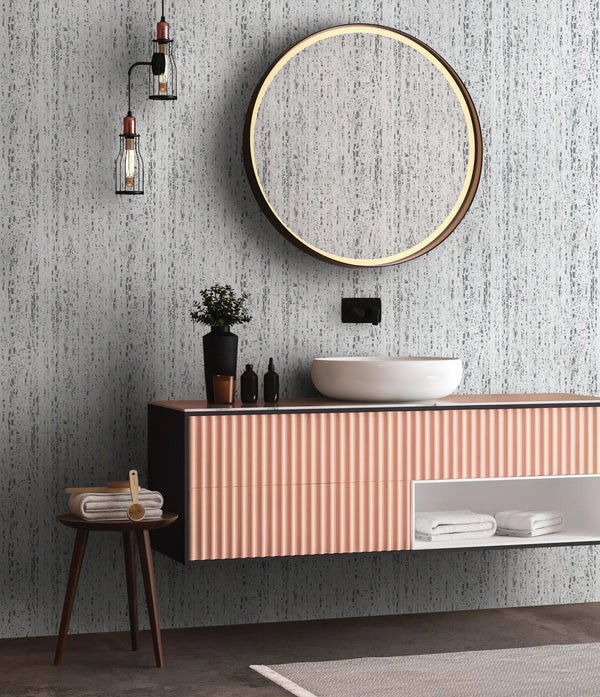 Marburg Travertino 33352 Gray and white vertically striated, stone-like wallpaper behind a bathroom setup with a round mirror, vessel sink on a floating vanity, pendant lights, and a small stool with folded towels.