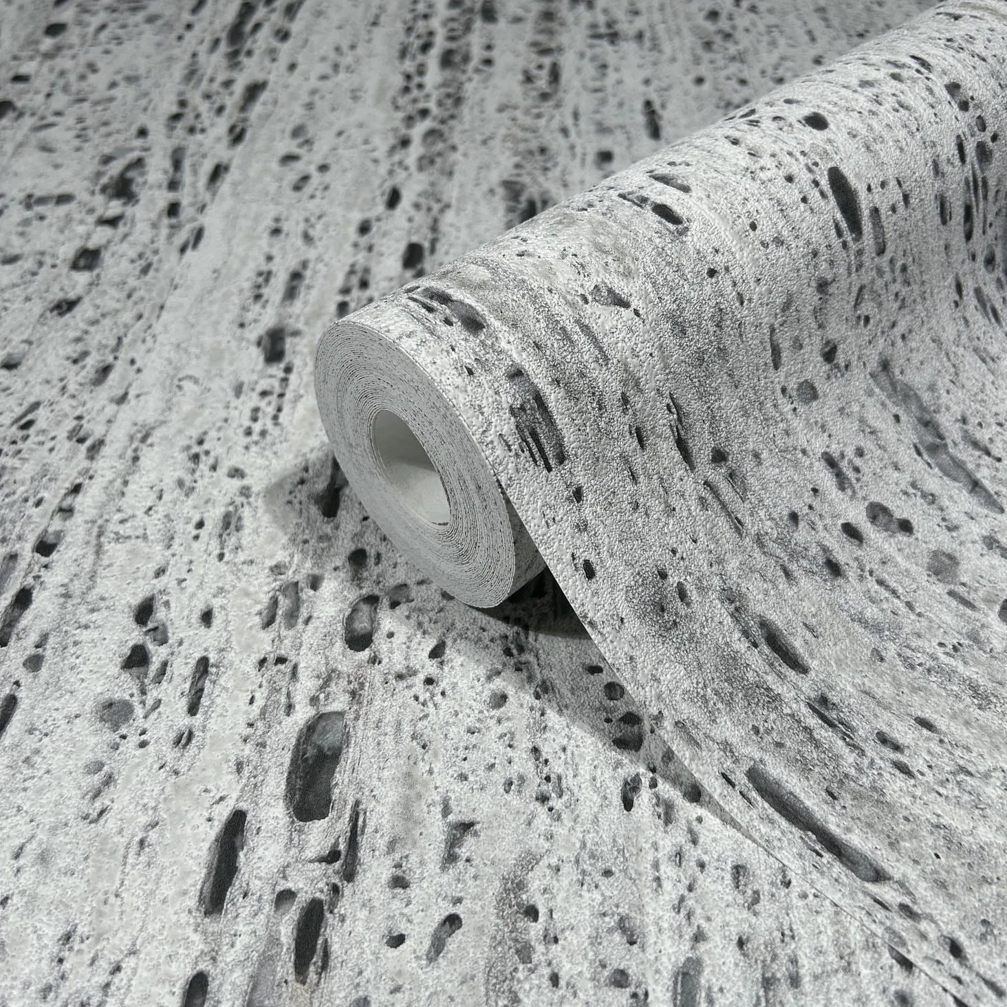 Marburg Travertino 33353 A wallpaper roll displayed on matching unrolled wallpaper, showing a gray travertine-style texture with irregular dark pits and speckles.