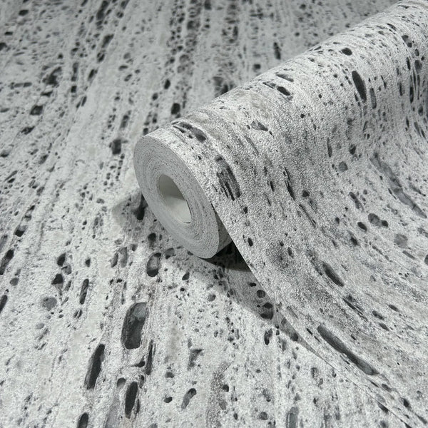 Marburg Travertino 33353 A wallpaper roll displayed on matching unrolled wallpaper, showing a gray travertine-style texture with irregular dark pits and speckles.