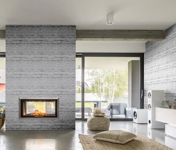 Marburg Travertino 33353 Modern living room featuring gray horizontally striated wallpaper around a built-in fireplace, with large sliding glass doors, a woven pouf, floor cushion, jute rug, white floor speakers, and a low media console.