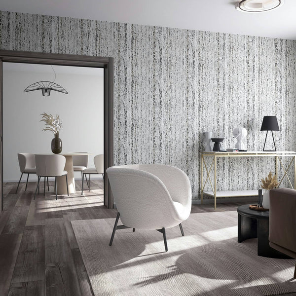 Marburg Travertino 33353 Living room scene featuring a light gray wallpaper with irregular vertical streaks and speckled texture. A white armchair, console table with a lamp and sculptural decor, round coffee table, and rug are in the foreground, with a doorway opening to a dining set.