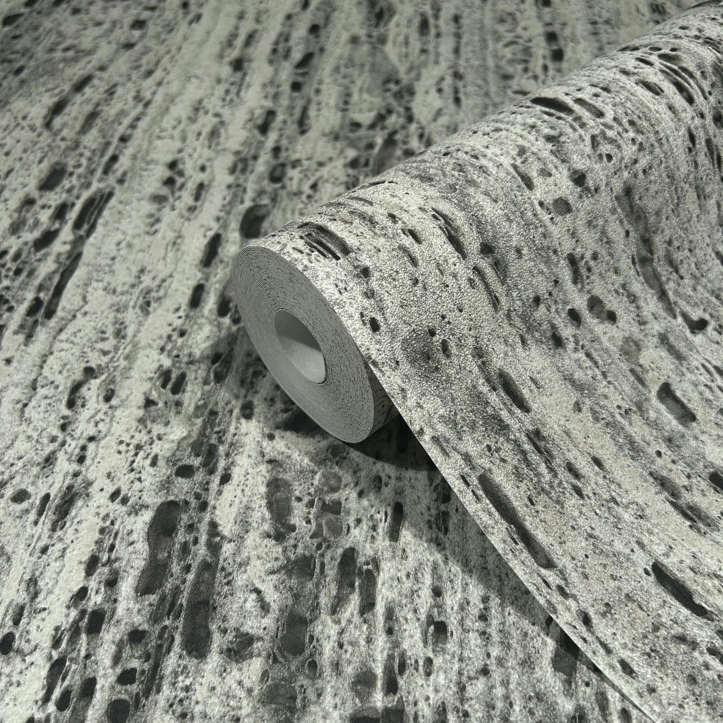 Marburg Travertino 33354 A wallpaper roll resting on matching unrolled wallpaper, showing a gray stone‑effect pattern with dark speckling and elongated pits.
