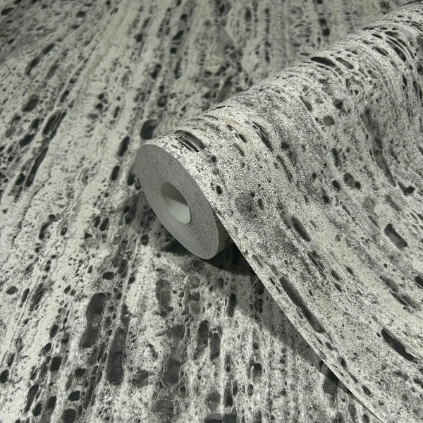 Marburg Travertino 33354 A wallpaper roll resting on matching unrolled wallpaper, showing a gray stone‑effect pattern with dark speckling and elongated pits.