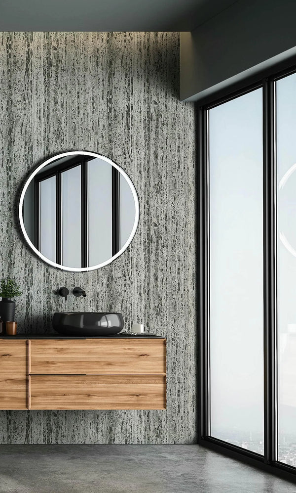 Marburg Travertino 33354 Gray stone-effect wallpaper with vertical striations shown in a bathroom behind a floating wood vanity with a dark vessel sink, round mirror, wall-mounted faucet, small plant, candle, and a large window.